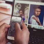 Pessoa segura um smartphone diante de um laptop que exibe imagens, em ambiente interno.