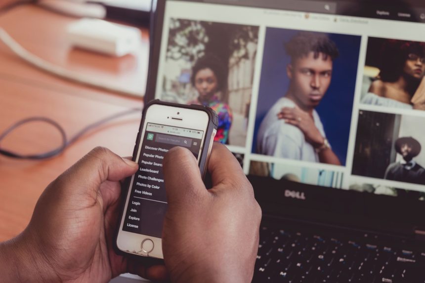 Pessoa segura um smartphone diante de um laptop que exibe imagens, em ambiente interno.