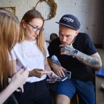 Três jovens conversam e usam dispositivos eletrônicos juntos em um café.