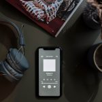 Smartphone exibindo um podcast ao lado de fones de ouvido e uma caneca sobre uma mesa.