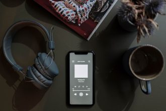 Smartphone exibindo um podcast ao lado de fones de ouvido e uma caneca sobre uma mesa.