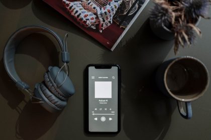 Smartphone exibindo um podcast ao lado de fones de ouvido e uma caneca sobre uma mesa.