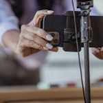Pessoa posiciona um smartphone em um tripé para gravar conteúdo, em ambiente interno desfocado.