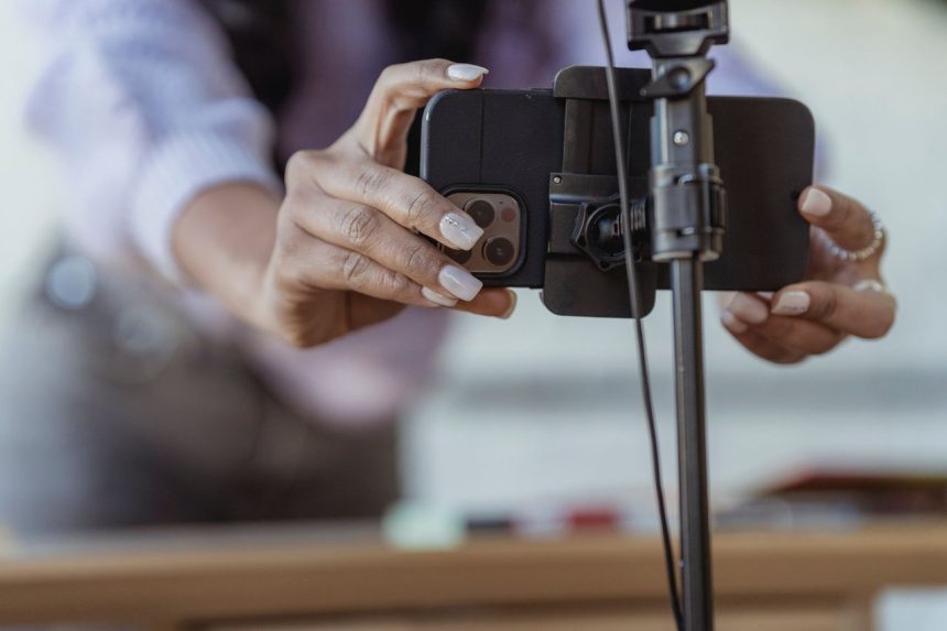 Pessoa posiciona um smartphone em um tripé para gravar conteúdo, em ambiente interno desfocado.