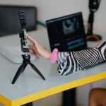 Criadora ajusta um smartphone em um tripé enquanto trabalha em um laptop, em um ambiente de produção e edição de vídeos.