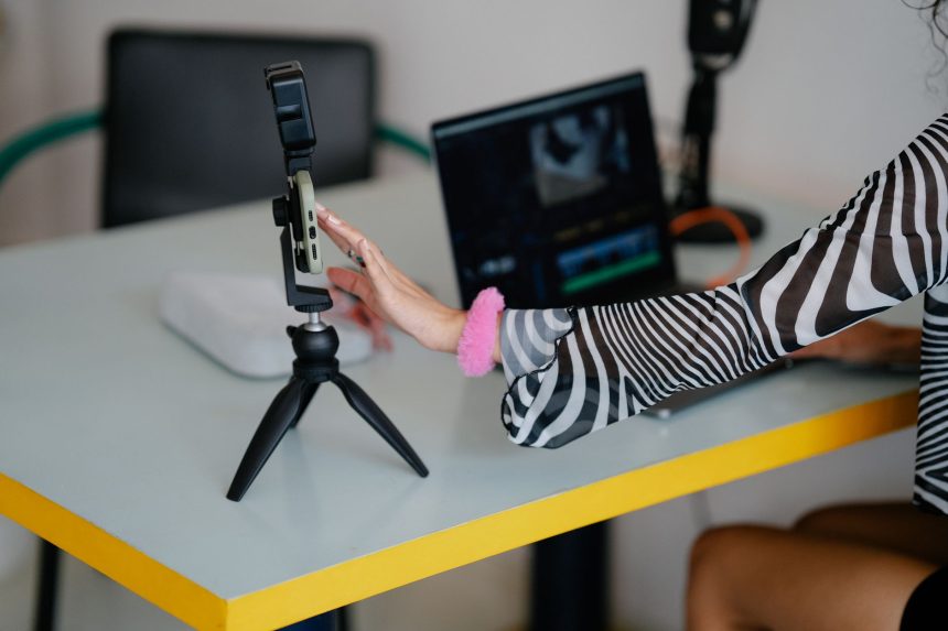 Criadora ajusta um smartphone em um tripé enquanto trabalha em um laptop, em um ambiente de produção e edição de vídeos.